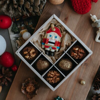 Chocolates natalinos numa caixa branca de chocolates junto a um papai noel de brinquedo, tudo sobre a mesa com decorações de natal